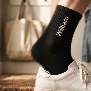 Op de afbeelding: Een zwarte geribbelde sok met de naam William in witte letters aan de zijkant, gedragen in een witte leren sneaker. De sok heeft een verticale ribbelstructuur en komt tot halverwege de kuit voor een strakke en eenvoudige uitstraling.
