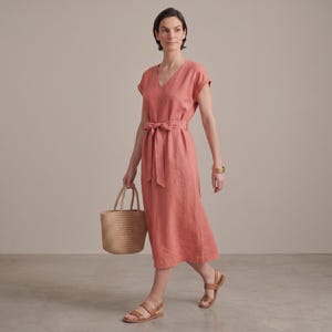 May include: A midi-length coral-colored dress with short sleeves, a V-neckline, and a matching fabric tie belt at the waist. The dress is paired with brown leather sandals and a woven straw tote bag, creating a cohesive and functional ensemble.