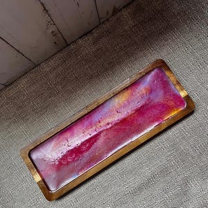 May include: A rectangular wooden tray featuring a smooth, glossy resin inlay. The resin surface displays a vibrant, swirling blend of deep magenta, bright pink, and soft lavender tones with subtle metallic gold highlights throughout.