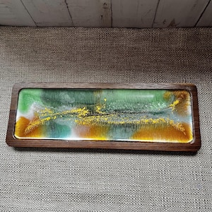 May include: A rectangular wooden tray featuring a smooth, glossy resin inlay. The resin surface displays a swirling abstract pattern of deep forest green, white, and metallic gold tones, with fine gold flakes scattered across the centre.