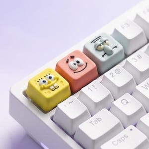 Peut inclure: Un clavier mécanique blanc doté de trois touches personnalisées sculptées. La touche jaune représente un personnage éponge souriant, la touche rose montre un personnage étoile de mer souriant et la touche bleu clair présente un personnage grincheux.