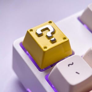 Peut inclure: Une touche de clavier mécanique carrée jaune avec un point d'interrogation argenté en relief sur la surface supérieure. La touche est placée sur une base de clavier blanche avec un rétroéclairage violet visible sous les touches.