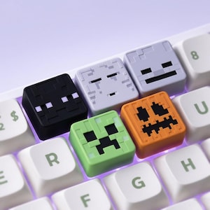 Peut inclure: Quatre touches de clavier carrées personnalisées représentant des visages de personnages pixélisés d'un jeu vidéo. L'ensemble comprend un enderman noir, un fantôme gris clair, un creeper vert et une citrouille orange sur un clavier blanc.