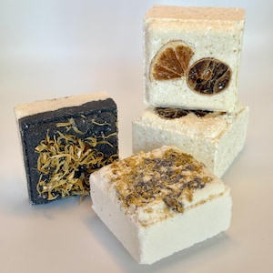 May include: A collection of four square bath bombs. One is black with yellow flower petals. Two are off-white with dried citrus slices embedded on the surface. One is off-white with a topping of dried purple and yellow flower petals.
