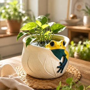 Op de afbeelding: Een witte keramische plantenpot in de vorm van een blad met een geel en donkergroen kikkerfiguurtje aan de rand. De pot bevat water en een kleine groene plant met brede bladeren, wat een decoratief vat vormt voor water- of bodemplanten.
