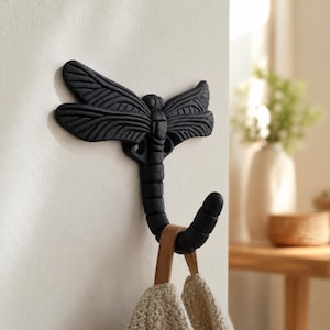 May include: A matte black wall hook shaped like a dragonfly with textured wings and a segmented body that curves upward to form a hook. A beige textured fabric item hangs from the lower hook section, displaying a neutral color palette.