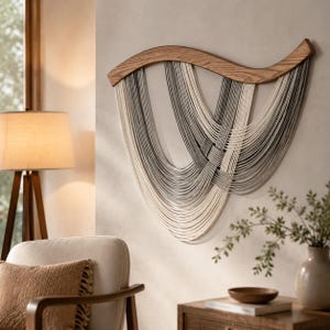 Può includere: Un arazzo da parete caratterizzato da un elemento superiore in legno curvato dai toni naturali, da cui pendono molteplici cordoncini di cotone nelle tonalità crema, grigio chiaro e grigio scuro, disposti a strati sovrapposti per un design fluido.