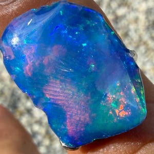 Könnte beinhalten: Ein roher, unpolierter Opal-Edelstein mit einem lebendigen Farbspiel in dominierenden tiefblauen Tönen, durchsetzt mit leuchtenden grünen, rosa und orangefarbenen Reflexen. Der Stein hat eine unregelmäßige, organische Form mit einer glatten Oberfläche.