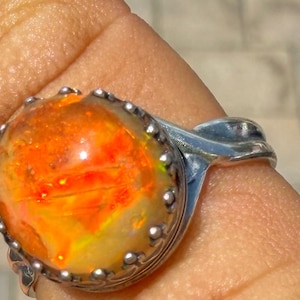 May include: A silver ring featuring a large, round, cabochon-cut opal gemstone. The opal displays vibrant flashes of orange, yellow, and red color play. The stone is secured in a scalloped, crown-like silver setting with a polished metal band.