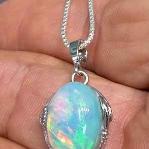 May include: A silver pendant necklace featuring an oval-shaped opal gemstone with iridescent flashes of blue, green, and pink. The opal is set in a polished silver frame with a beaded edge, suspended from a delicate silver box chain necklace.