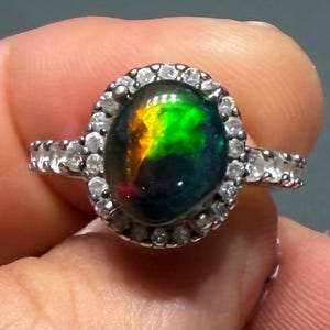 May include: A silver ring featuring a central oval-shaped black opal with vibrant green and orange flashes. The gemstone is surrounded by a halo of small, clear, round-cut stones, with additional clear stones set along the band of the ring.