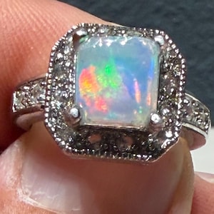 Könnte beinhalten: Ein silberfarbener Ring mit einem rechteckigen, irisierenden Opal in der Mitte, der grüne, orangefarbene und blaue Farbspiele zeigt. Der Opal ist von einem Kranz aus kleinen, klaren, funkelnden Steinen in einer quadratischen Fassung umgeben.