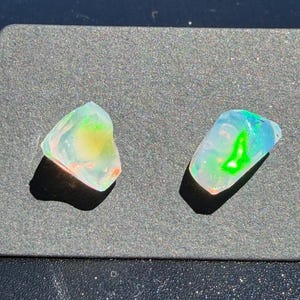 May include: Two raw, translucent opal gemstones with irregular shapes. The stones exhibit a play of color, featuring vibrant flashes of neon green, yellow, and orange against a milky, semi-transparent base. Each stone has a smooth, polished surface.