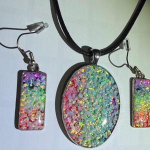 May include: A jewelry set consisting of an oval pendant on a black cord necklace and two matching rectangular drop earrings. Each piece features a vibrant, multi-colored crushed stone inlay with iridescent pink, green, and blue flecks.