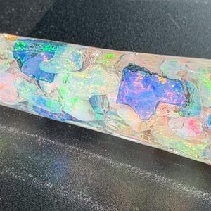 May include: A cylindrical resin blank embedded with numerous iridescent opal fragments. The material is clear and polished, showcasing a variety of colorful stone pieces in shades of blue, green, pink, and white that reflect light with a vibrant, multi-colored shimmer throughout the entire structure.