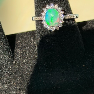 Könnte beinhalten: Ein silberfarbener Ring mit einem zentralen, ovalen Opal, der lebhafte grüne und rote Blitze zeigt. Der Opal ist von einem Kranz aus kleinen, klaren, runden Steinen umgeben, und die Ringschiene ist seitlich mit weiteren kleinen Steinen besetzt.
