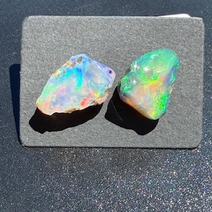 May include: Two raw, unpolished opals with an iridescent play of color. The left opal displays flashes of blue, green, and orange against a translucent white base. The right opal features vibrant green and orange hues with a smooth, rounded shape.