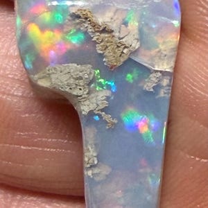 Könnte beinhalten: Ein Anhänger mit einem polierten, unregelmäßig geformten Opal mit einer durchscheinenden, milchig blauen Basis. Der Stein zeigt lebhafte grüne, gelbe und rote Feuerblitze, durchsetzt mit strukturierten, hellen natürlichen Matrixeinschlüssen.