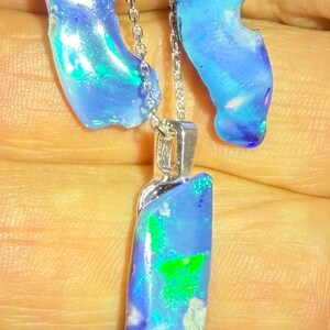 Könnte beinhalten: Ein Schmuckset bestehend aus einem Anhänger an einer Silberkette und zwei passenden Ohrringen. Jedes Stück zeigt einen polierten, unregelmäßigen blauen Opal mit leuchtend grünen und irisierenden Reflexen, gefasst in glänzendem Silber.