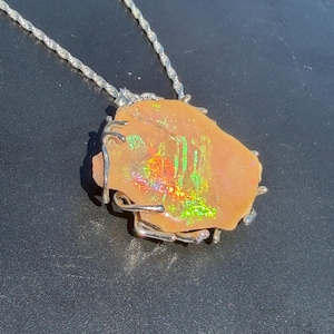 Könnte beinhalten: Eine Anhängerkette mit einem rohen, pfirsichfarbenen Opalstein, der lebhafte grüne und rote irisierende Blitze aufweist. Der Stein ist in einer silberfarbenen Metalldrahtfassung befestigt und hängt an einer passenden gedrehten Kette.