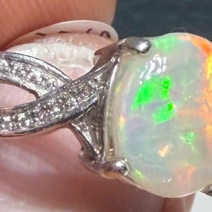 Könnte beinhalten: Ein silberfarbener Ring mit einem großen, runden, irisierenden Opal-Cabochon, der lebhafte grüne und orangefarbene Lichtreflexe aufweist. Die Ringschiene hat ein geteiltes, überkreuztes Design, das mit kleinen, klaren Steinen besetzt ist.
