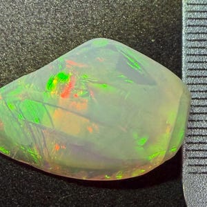 May include: A translucent, irregularly shaped opal gemstone displaying vibrant flashes of green and orange play-of-colour. The stone is positioned next to a metal ruler, indicating a length of approximately 1.5 centimetres at its widest point.
