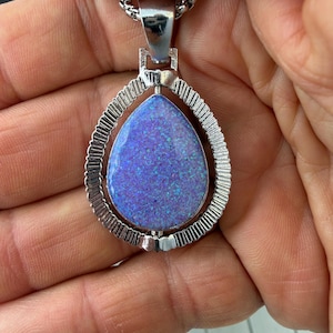 May include: A teardrop-shaped pendant featuring a vibrant purple and blue iridescent stone set within a textured silver-toned metal frame. The pendant is attached to a silver-toned chain, showcasing a polished finish and a detailed, ridged border.