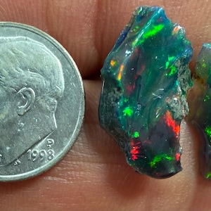 Könnte beinhalten: Zwei rohe, dunkel getönte Opale mit lebhaften grünen, blauen und roten Farbspielen, platziert neben einer amerikanischen Zehn-Cent-Münze als Maßstab, die einen Durchmesser von etwa 1,79 cm hat und einen klaren Größenvergleich bietet.