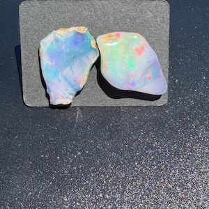 Könnte beinhalten: Zwei rohe, unregelmäßige Opal-Edelsteine mit einer durchscheinenden weißen Basis und lebhaften Farbspielen in Blau, Grün und Rot. Die Steine haben natürliche, unpolierte Kanten und sind nebeneinander auf einer rechteckigen schwarzen Karte angeordnet.