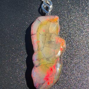 Könnte beinhalten: Ein Anhänger mit einem unregelmäßigen, durchscheinenden Opal-Edelstein mit lebhaften roten, gelben und grünen Farbspielen, der an einer silberfarbenen Metallkette mit einer dekorativen, gedrehten Metallöse befestigt ist.