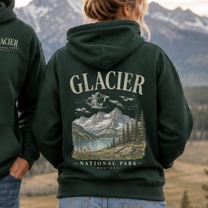 Pode incluir: Uma camisola com capuz verde-escura com um pequeno log&oacute;tipo do Glacier National Park no peito e um gr&aacute;fico grande e detalhado nas costas que retrata uma cordilheira, um lago e pinheiros com o texto Glacier National Park Montana.