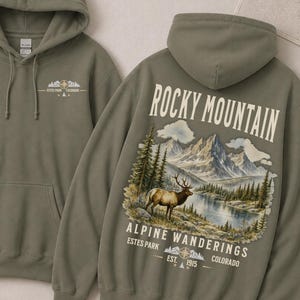 May include: An olive green hooded sweatshirt featuring a small graphic on the front chest and a large back print depicting a mountain landscape with an elk, pine trees, a lake, and the text Rocky Mountain Alpine Wanderings Estes Park Colorado.