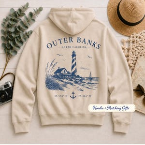 Pu&ograve; includere: Una felpa con cappuccio color beige con una grafica blu scuro sul retro. Il design include un faro, una piccola casa, una barca a vela, il testo Outer Banks North Carolina, le coordinate 35.558 N 75.466 W e un'icona a forma di ancora.