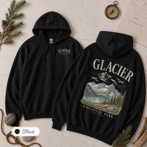 Pode incluir: Uma camisola com capuz preta com um pequeno log&oacute;tipo do Glacier National Park no peito esquerdo e um grande gr&aacute;fico nas costas que retrata uma paisagem montanhosa com um lago, pinheiros e o texto Glacier National Park Montana.