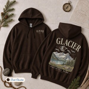 Pode incluir: Uma camisola com capuz castanho-escuro com um bolso frontal e capuz com cord&otilde;es. O peito apresenta o texto Glacier National Park. As costas exibem um gr&aacute;fico de montanhas, um lago e &aacute;rvores com o texto Glacier National Park Montana.