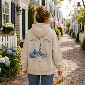 Pu&ograve; includere: Una felpa con cappuccio color beige con una grafica blu sul retro. Il design include un faro, una piccola casa, erba alta, uccelli, una barca a vela, un'ancora e il testo Outer Banks North Carolina con le coordinate 35.558 N 75.466 W.