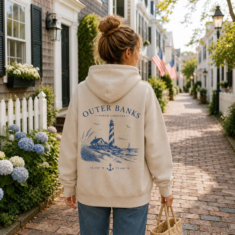 Pu&ograve; includere: Una felpa con cappuccio color beige con una grafica blu sul retro. Il design include un faro, una piccola casa, erba alta, uccelli, una barca a vela, un'ancora e il testo Outer Banks North Carolina con le coordinate 35.558 N 75.466 W.
