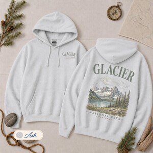 Pode incluir: Uma camisola com capuz cinzento-claro com um pequeno log&oacute;tipo do Glacier National Park no peito esquerdo e um grande gr&aacute;fico nas costas que mostra uma cordilheira, um lago e pinheiros com o texto Glacier National Park Montana.