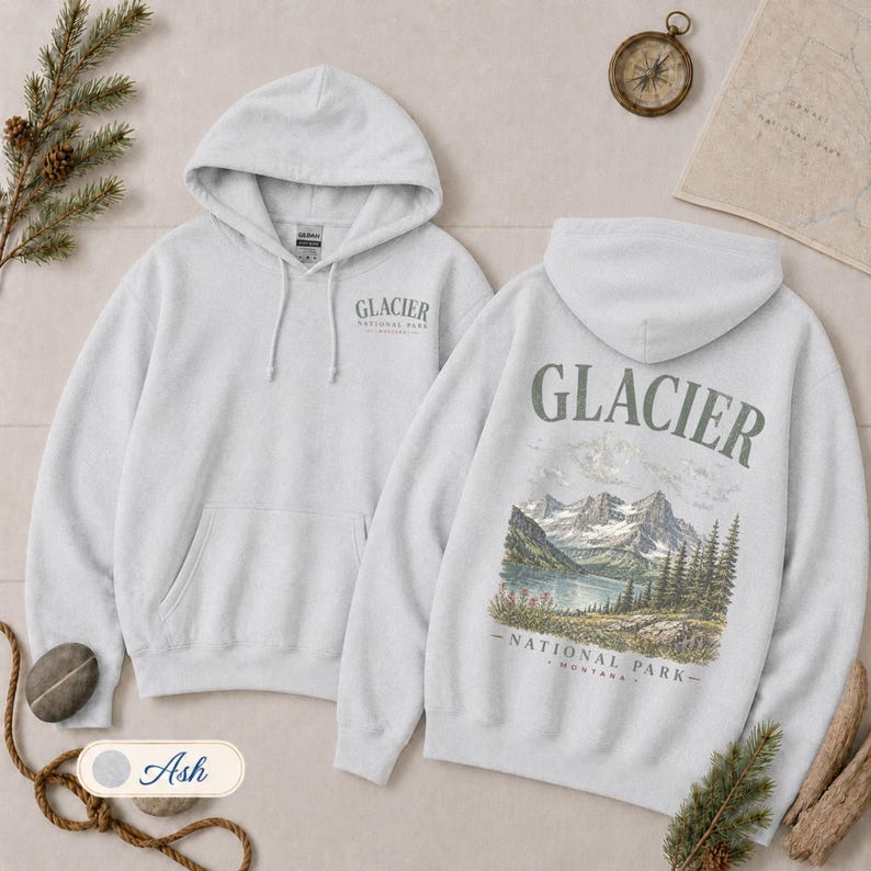 Pode incluir: Uma camisola com capuz cinzento-claro com um pequeno log&oacute;tipo do Glacier National Park no peito esquerdo e um grande gr&aacute;fico nas costas que mostra uma cordilheira, um lago e pinheiros com o texto Glacier National Park Montana.
