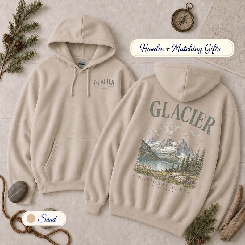 Pode incluir: Uma camisola com capuz cor de areia com um pequeno log&oacute;tipo do Glacier National Park no peito esquerdo e um gr&aacute;fico grande e detalhado nas costas que mostra uma cordilheira, um lago e pinheiros com o texto Glacier National Park Montana.