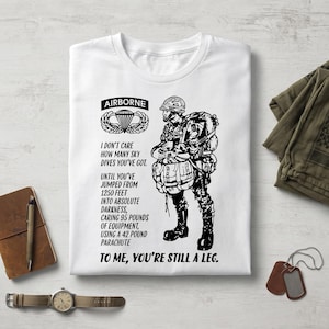 Peut inclure: Un t-shirt blanc avec une illustration noire d'un parachutiste équipé. Le texte indique : AIRBORNE, I DON'T CARE HOW MANY SKY DIVES YOU'VE GOT. UNTIL YOU'VE JUMPED FROM 1250 FEET INTO ABSOLUTE DARKNESS, CARING 95 POUNDS OF EQUIPMENT, USING A 42 POUND PARACHUTE. TO ME, YOU'RE STILL A LEG.