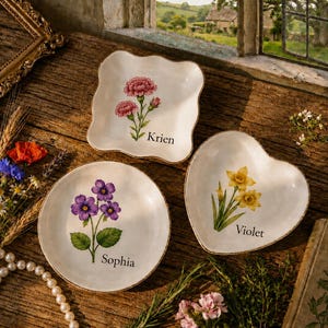 May include: Three ceramic trinket dishes with scalloped, round, and heart-shaped edges. Each white dish features a botanical illustration of flowers and a name: pink carnations labelled Krien, purple violets labelled Sophia, and yellow daffodils labelled Violet.