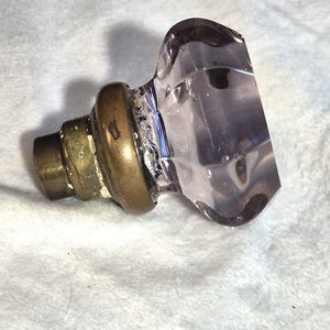 Puede incluir: Un pomo de cristal facetado de color morado claro con un acabado transparente y translúcido. El pomo está unido a una base metálica circular de tono latón desgastado con un poste de montaje cilíndrico que sobresale del centro de la pieza.