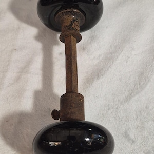 May include: A vintage door knob set featuring two smooth, round, glossy black knobs connected by a central square metal spindle. The metal components show visible signs of oxidation and rust, including a small set screw on the neck of one knob.