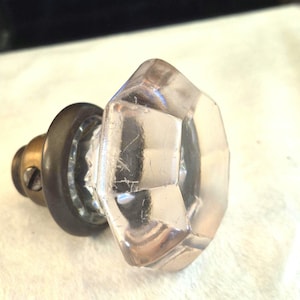 Peut inclure: Un bouton de placard en verre transparent à facettes de forme octogonale. Le bouton présente une base circulaire métallique sombre avec un mécanisme de vis visible sur le côté, reliant la tête en verre transparent à la fixation.