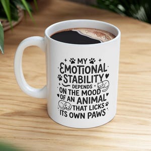 Puede incluir: Una taza de cerámica blanca llena de café negro, con texto en negro que dice: MY EMOTIONAL STABILITY DEPENDS ON THE MOOD OF AN ANIMAL THAT LICKS ITS OWN PAWS, junto a pequeñas ilustraciones de huellas, corazones, un gato y un perro.