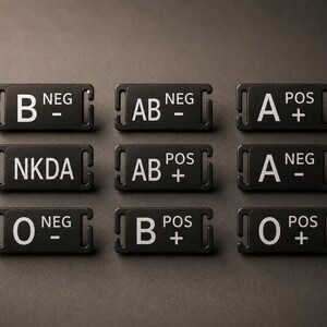 Peut inclure: Un ensemble de neuf étiquettes d'identification médicale rectangulaires noires avec du texte blanc. Chaque étiquette indique un groupe sanguin spécifique, dont B NEG, AB NEG, A POS, NKDA, AB POS, A NEG, O NEG, B POS et O POS, avec des fentes latérales.