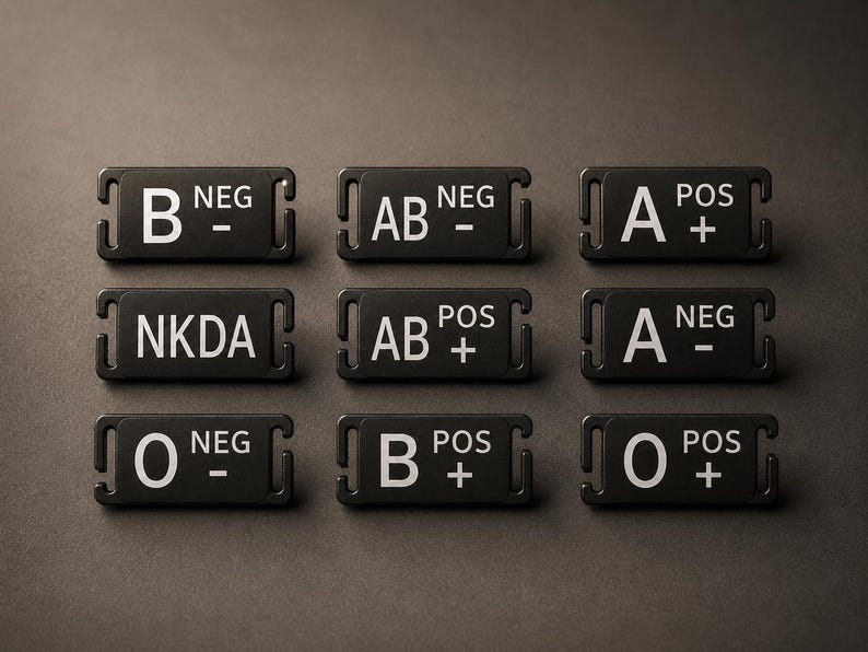 May include: A set of nine rectangular black medical identification tags with white text. Each tag features a specific blood type, including B NEG, AB NEG, A POS, NKDA, AB POS, A NEG, O NEG, B POS, and O POS, with integrated side slots for attachment.