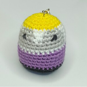 Puede incluir: Un pequeño llavero de peluche tejido a ganchillo, de forma redonda, con rayas horizontales de hilo amarillo, blanco, gris claro y morado. El objeto tiene dos ojos pequeños, oscuros y bordados, y una argolla metálica plateada en la parte superior.