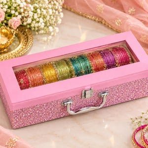 May include: A rectangular pink glitter storage box with a clear glass lid, featuring a silver metal latch and handle. Inside, a row of multi-coloured bangles in shades of red, orange, yellow, green, teal, and purple are arranged in a line.